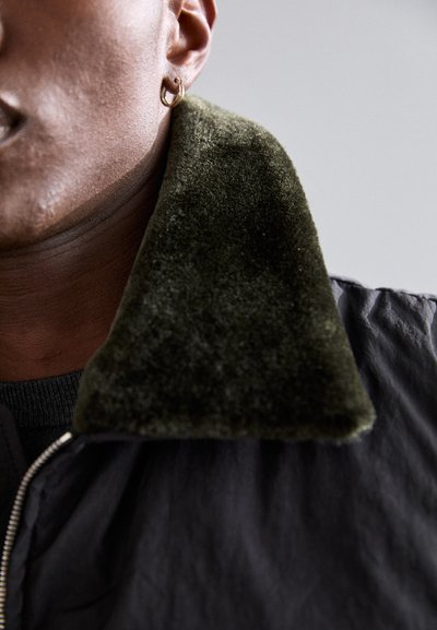 Črna jakna z teksturiranim, temno zelenim žametnim ovratnikom. Prižgana slika, ki prikazuje del vratu in uhans. Previden sijaj na blagu.