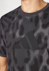 Melnas sporta T-krekls ar smalku pelēku leoparda rakstu, apaļu izgriezumu un lielu melnu Adidas logotipu priekšpusē.
