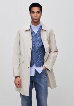 Mężczyzna w beżowym płaszczu, niebieskiej pikowanej kamizelce, jasnoniebieskiej koszuli i niebieskich dżinsach, stojący na tle jednolitego jasnego tła.