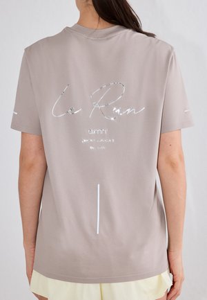 Osoba o długich ciemnych włosach nosi jasnoszary T-shirt z błyszczącym srebrnym napisem "Le Run" i logo marki na plecach, w połączeniu z jasnymi szortami.