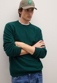 Grüner Wollpullover mit gerippten Bündchen, Rundhalsausschnitt und lockerem Schnitt. Kombiniert mit blauen Jeans. Braune Fischgrätmütze mit grünem Logoakzent.