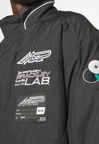 Svart vadderad jacka med vita broderade logotyper och etiketter, hög krage och en texturerad yta. Accenter inkluderar färgade cirkulära lappar.