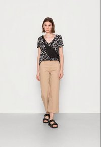 Top com padrão floral em preto, calças largas em bege e sandálias pretas com tiras grossas. Mala de crossbody usada ao ombro. Fundo simples.