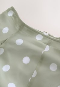 Tissu vert clair avec de gros pois blancs, montrant des coutures cousues et une fermeture éclair cachée le long du bord.
