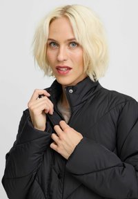 Černá péřová bunda s prošívaným designem, vysokým límcem a zipem; má akcenty na cvoky a jemnou texturu.