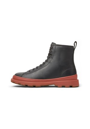Bota negra de cuero con cordones y suela roja gruesa y texturizada, con una pestaña en el talón, vista de lado sobre un fondo blanco.
