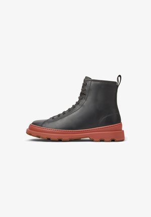 Bottines en cuir noir à lacets avec une semelle rouge épaisse et texturée et une languette à l'arrière, vue de côté sur un fond blanc.