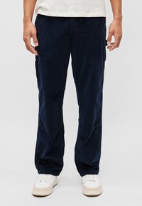 Pantaloni din corduroy bleumarin cu o croială dreaptă, având un material texturat, buzunare laterale și încălțăminte albă cu accente bej.