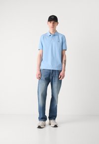 Lys blå polo skjorte, blå jeans, grå joggesko. Svart caps med hvitt logo. Person står på en ensfarget bakgrunn.