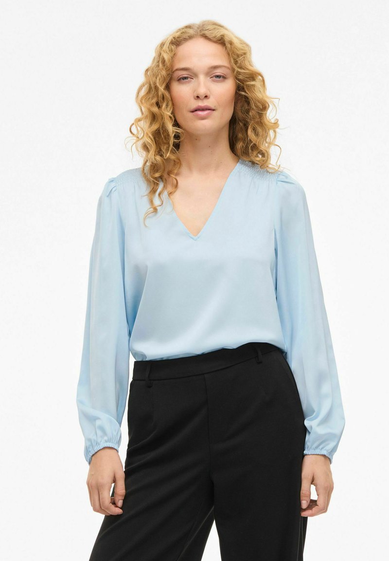 Blouse bleu clair en tissu soyeux avec un col en V et des manches longues aux poignets froncés, associée à un pantalon noir ajusté.