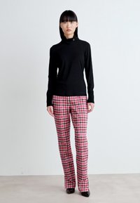 Schwarzes langärmliges Oberteil mit hohem Kragen und seitlichen Knopfdetails, kombiniert mit rot-schwarz karierter Hose. Glatte Textur.