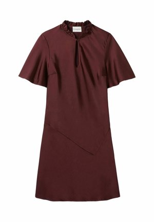 Femme debout portant une veste en jean bleue avec des boutons dorés sur une robe bordeaux et des mocassins noirs, sur un fond clair uni.