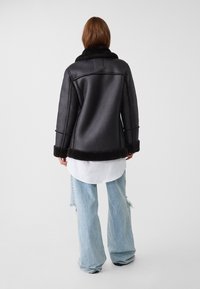 Kvinna klädd i svart lammulljacka med pälskrage och manschetter, vit skjorta och ljusblå slitna vida jeans, stående på en neutral bakgrund.