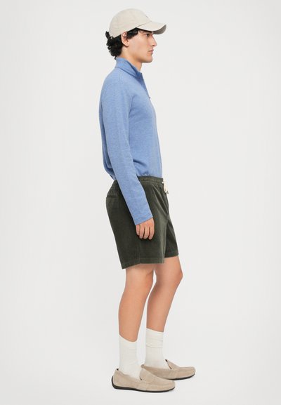 Camisa azul de mangas longas, calções verde-azeitona, meias brancas e sapatilhas bejes de calçar. Apresenta colarinho e cintura com cordão ajustável.