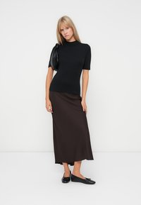 Schwarzes kurzärmliges Oberteil und brafter Satin-Midirock-Outfit. Das Model hält eine schwarze Handtasche und trägt schwarze Slippers.