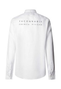 Camisa blanca de manga larga hecha de lino. Presenta un cuello clásico y botones en los puños. La parte posterior muestra el texto en azul marino, "FAÇONNABLE FRENCH RIVIERA."
