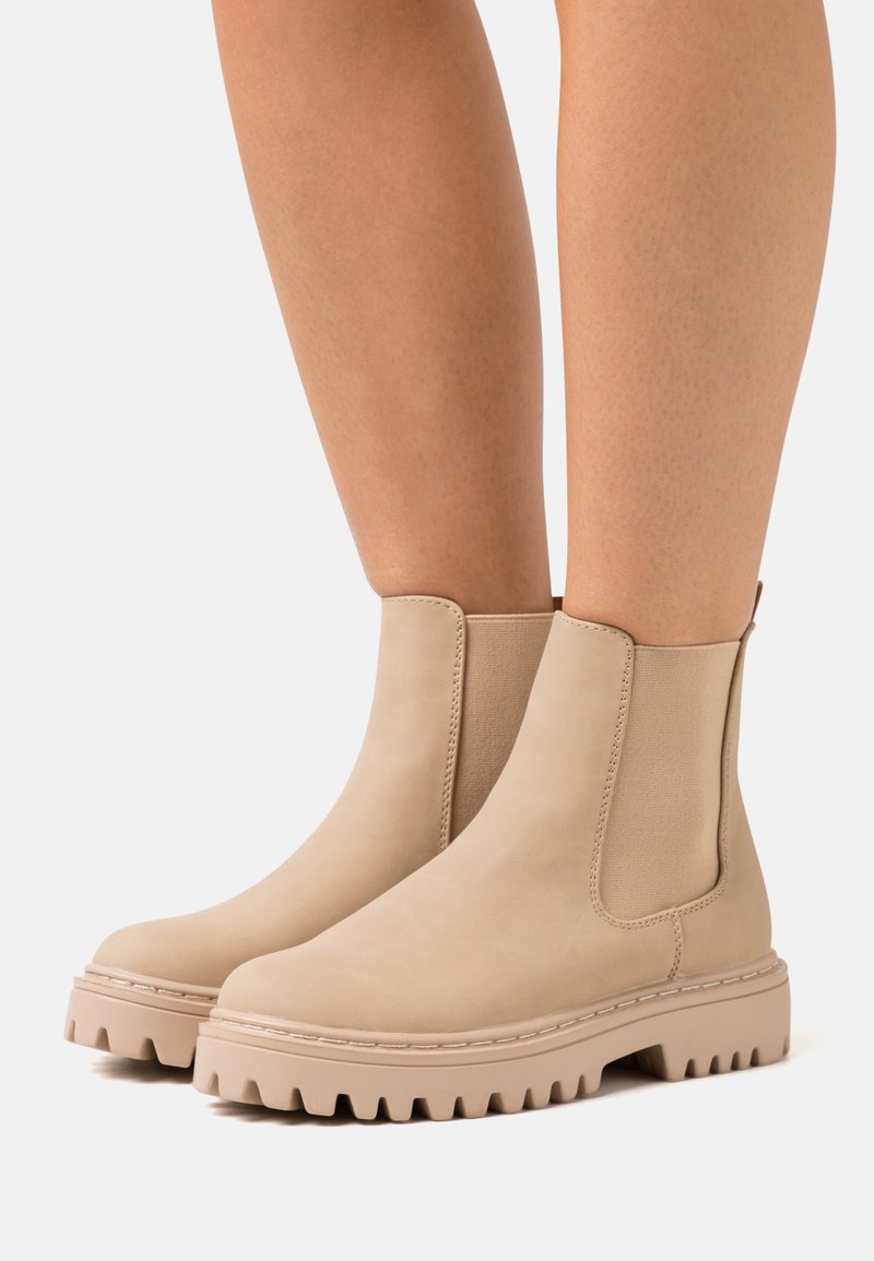 Vue en longueur de jambe de bottines beiges à semelle épaisse portées sur jambes nues, montrant des panneaux latéraux élastiques et des languettes de tirage sur un fond clair.