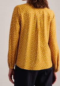Yellow blouse with white leaf pattern, long sleeves, and a rounded hem. Lightweight fabric with a smooth texture, buttoned cuff details.