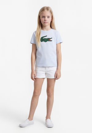 Fiatal lány hosszú szőke hajjal, világoskék pólóban zöld krokodil grafikával, fehér rövidnadrágban és fehér bebújós cipőben áll.