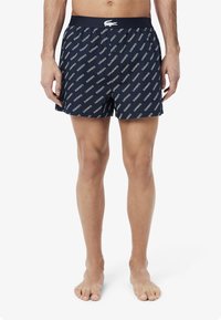 Navy swim shorts featuring an all-over white logo pattern, elastic waistband, and a relaxed fit. Made from lightweight, breathable fabric.