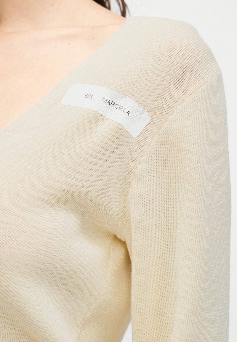 Pull en maille couleur crème avec col en V et finition texturée. Présente une étiquette rectangulaire avec l'inscription "SIX MARGIELA" imprimée en noir.