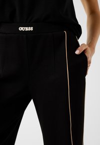 Nahaufnahme von schwarzen Hosen mit beiger Seitenpaspelierung, einer "GUESS"-Logoschnalle und einer Hand, die in der rechten Tasche ruht.