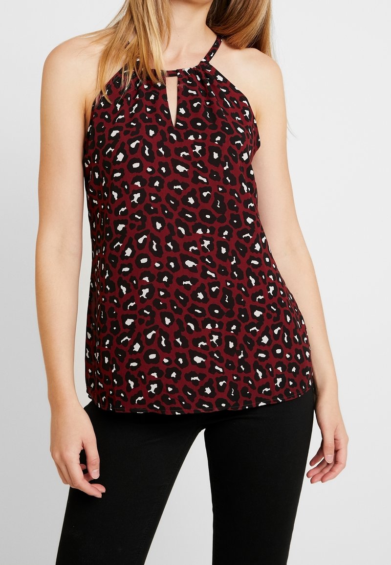 Femme portant un haut sans manches bordeaux avec imprimé léopard noir et blanc et encolure en forme de goutte, associée à un pantalon noir.