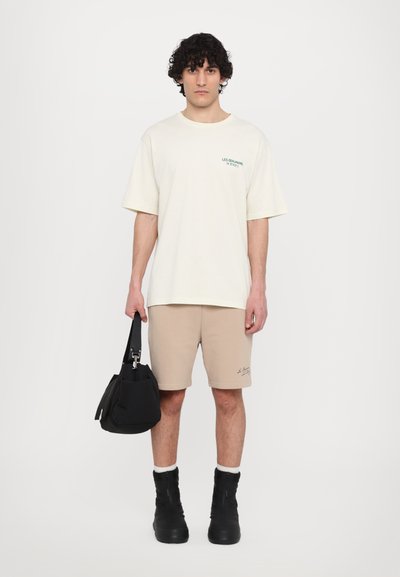 T-shirt beige clair avec texte vert, associé à un short beige. Le mannequin porte un sac noir. Des bottes isolées noires complètent la tenue.