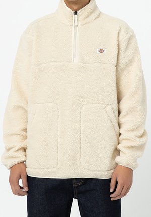 Man wearing off-white fleece pullover with quarter zip, two front pockets, and small logo on chest, paired with dark blue jeans.