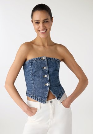 Donna sorridente che indossa un top di jeans blu senza spalline con bottoni frontali e pantaloni bianchi, con le mani nelle tasche, su uno sfondo semplice.