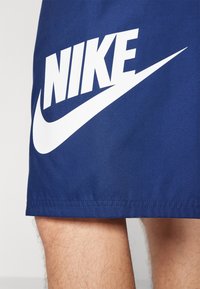 Niebieskie spodenki sportowe wykonane z gładkiej tkaniny, z dużym białym logo „NIKE” i zakrzywionym wzorem Swoosh.