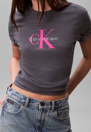 Osoba nosząca dopasowany ciemnoszary t-shirt Calvin Klein z różowym logo oraz jasnoniebieskie dżinsy z wysokim stanem z metką Calvin Klein.