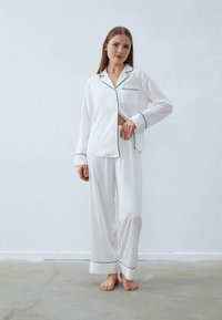 Vitt pyjamaset i mjukt tyg. Långärmad topp med svart kant och bröstficka. Löst sittande byxor med vida ben och fållade nederdelar.