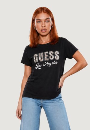 Mujer con cabello rojo que lleva una camiseta negra de Guess Los Ángeles y vaqueros azul claro, de pie con una mano en la cadera.