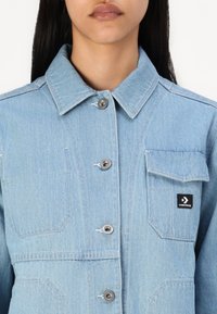 Lyseblå denimjakke med knaplukning, spidse krave og en brystlomme med et sort Converse-logo-patch.