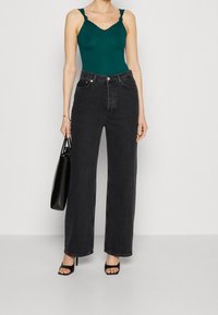 Grön bodysuit med knutna axelband, i kombination med högmidjade, vida svarta jeans. Modellen håller en svart väska med texturerad yta.