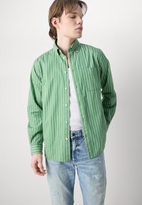 Πράσινο ριγέ πουκάμισο με κουμπιά, κατασκευασμένο από βαμβάκι, με μακριά μανίκια και τσέπη στην αριστερή πλευρά, συνδυασμένο με φθαρμένα ανοιχτόχρωμα μπλε τζιν.