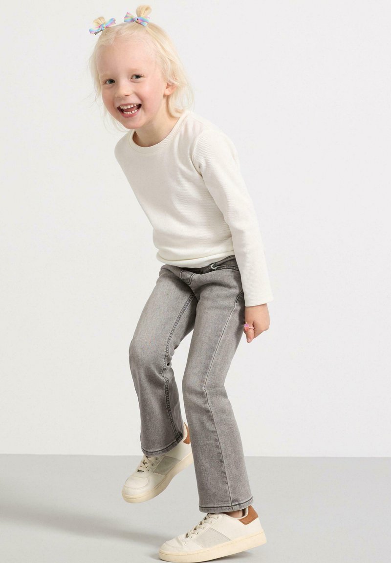 Ljusbeige långärmad topp, grå vidbyxor och vita sneakers med bruna detaljer. Barn som hoppar med två små fl bows i blont hår.