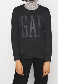 Sudadera negra con un gran logo "GAP" brillante en el frente. Fabricada con una tela suave, tiene mangas largas y un cuello redondo.
