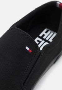 Černé nazouvací boty s textilním svrškem, šitými detaily a malou logo nášivkou v červené, bílé a modré barvě na boku. Plochá podrážka, casual design.
