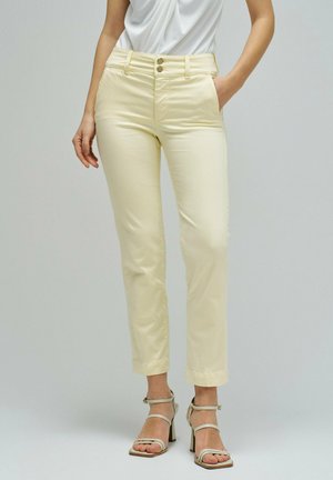 Femme portant un pantalon crème taille haute avec fermeture à double bouton et des sandales à talons beige, debout avec une main dans la poche.