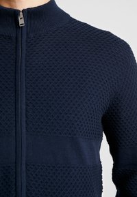 Marintröja med dragkedja som har ett texturerat våffelstickat mönster med släta sektioner på torso och ärmar. Hög krage med en metallisk dragkedja.