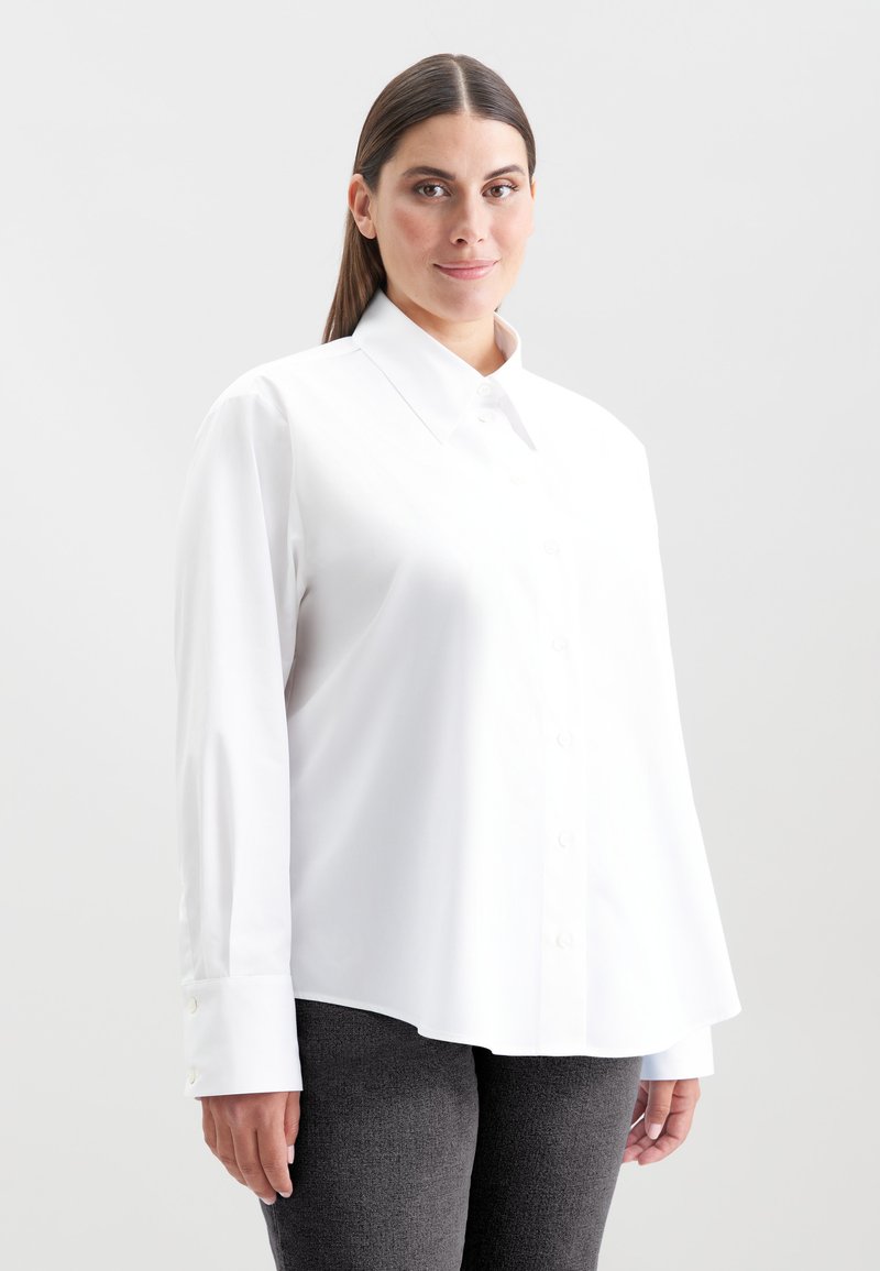 Donna con lunghi capelli lisci, che indossa una camicia bianca con bottoni e colletto ampio e pantaloni grigio scuro, in piedi contro uno sfondo grigio chiaro.