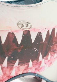 Close-up of a shoe with a graphic design featuring sharp white shark teeth and a metallic logo emblem on the upper surface.