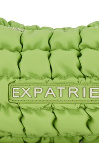 Geantă din piele verde cu textură matlasată și închidere cu fermoar. Prezintă un logo „EXPATRIÉ” proeminent în bej pe o etichetă din piele asortată.