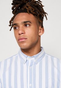 Camisa abotonada con rayas verticales en azul claro y blanco, de tela de algodón, con cuello doblado y un pequeño logo en el pecho.