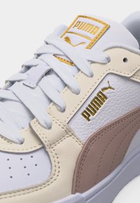 Κοντινό πλάνο λευκού αθλητικού παπουτσιού Puma με μπεζ και καφέ λεπτομέρειες, λευκά κορδόνια και χρυσό λογότυπο Puma στη πλευρά και στη γλώσσα του παπουτσιού.