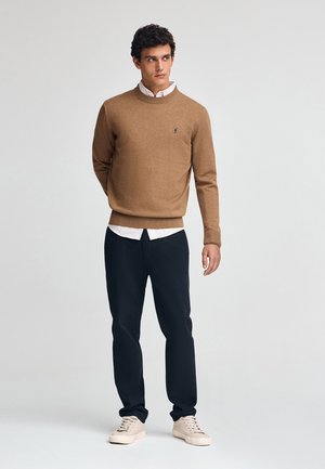 Bruine gebreide trui met ronde halslijn over een witte geknoopte blouse, gecombineerd met donkere broeken en lichtgekleurde sneakers. Eenvoudig ontwerp, zonder patronen.