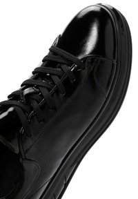 Deportivas de cuero negro con acabado brillante, con cordones negros y una suela de goma texturizada. Punta redondeada y elementos de diseño minimalista.