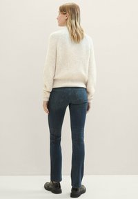 Suéter de crema grueso con textura acanalada, combinado con jeans ajustados azul oscuro y zapatos planos negros, visto desde atrás.